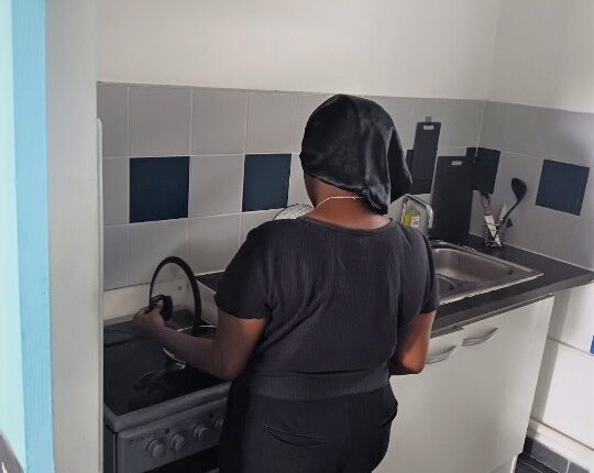 Une jeune femme est visible de dos, en train de soulever le couvercle d'une casserole dans la cuisine d'un appartement